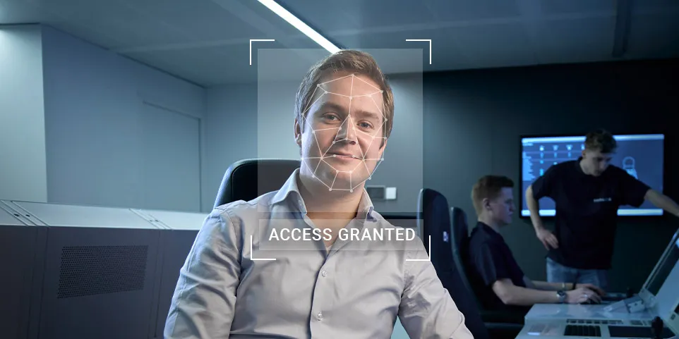 Man working at a bank of screens with biometric tech overlay on his face and the text 'access granted' below